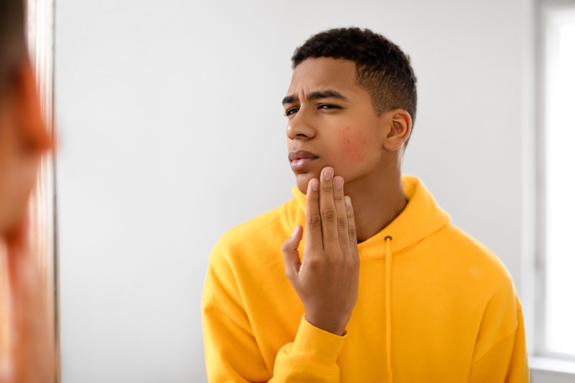 Concerned black teen guy examining face in the mirror